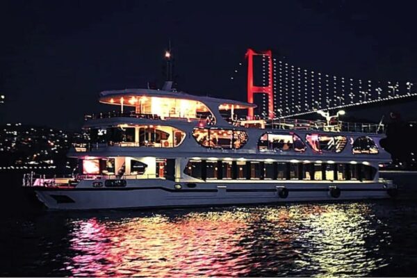 Luxury dinner cruise ship on the Bosphorus at night, illuminated with warm lights, with the Bosphorus Bridge in the background.