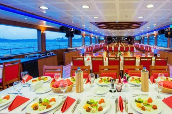 Elegant dinner cruise setup on a boat with large windows, red chairs, and views of the sea at sunset, offering fine dining ambiance.